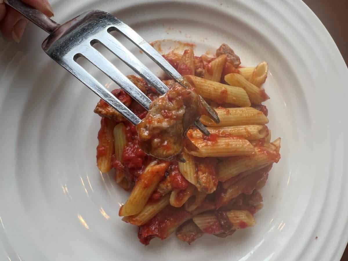 Top down image of a fork with Italian Sausage on it hovering over a plate of penne.