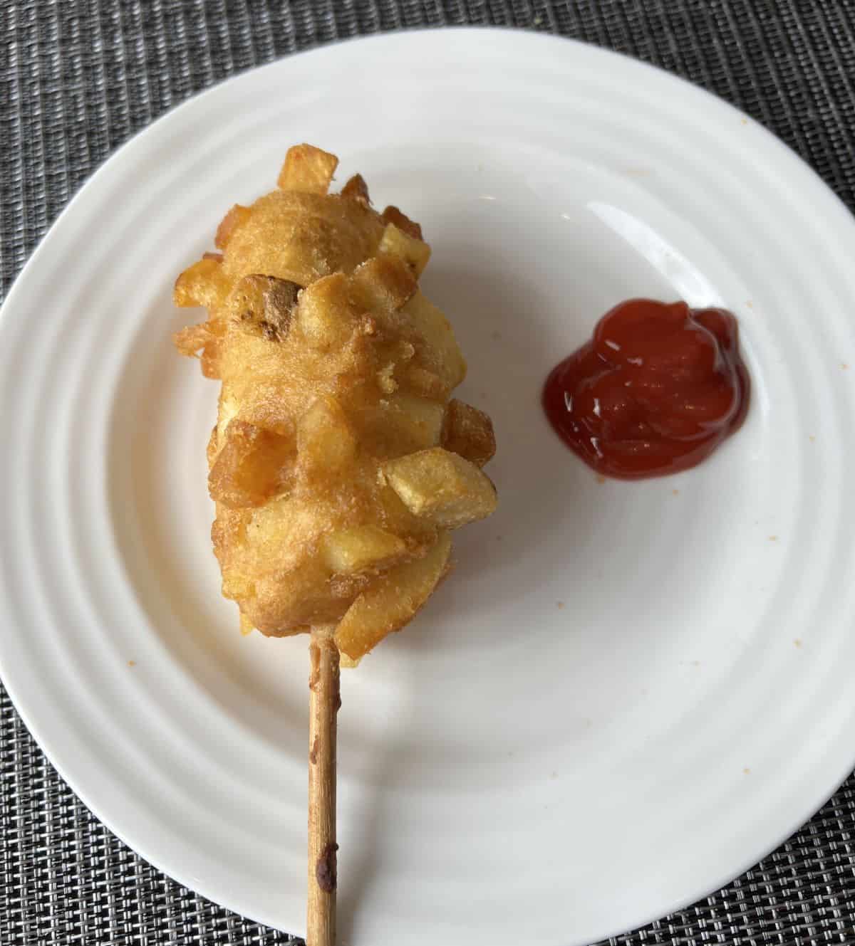 Top down image of a crispy potato corn dog served on a white plate with a side of ketchup. 