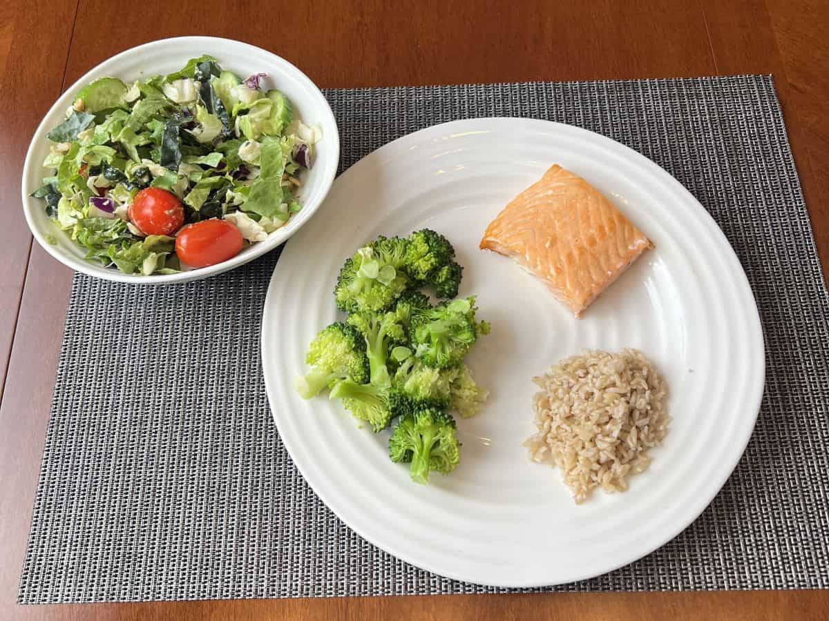 Image of salmon served on plate beside a bed of rice and broccoli, beside the plate is a salad.  