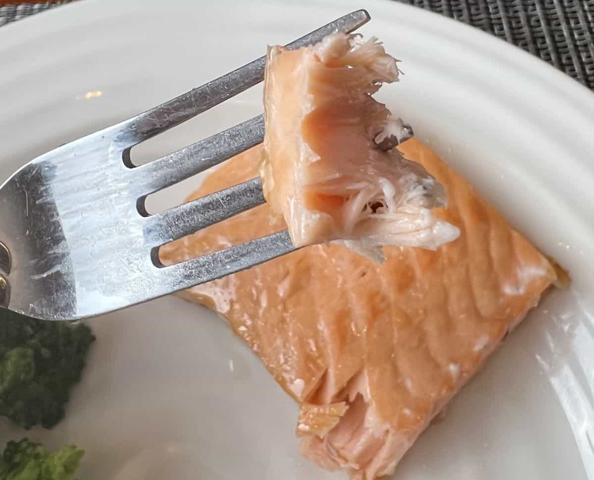 Closeup image of a fork with a flaky moist bite of salmon on it hovering over a filet of salmon.