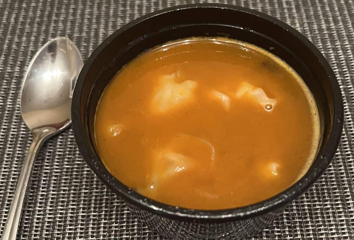 Top down image of the bowl of shrimp wonton soup in tom yum base sitting on a table beside a spoon.