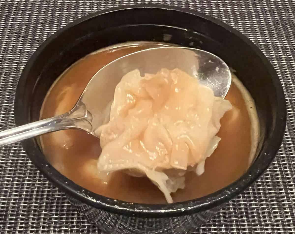 Image of a spoon with a shrimp wonton on it hovering over a bowl of tom yum broth with wontons in the broth.