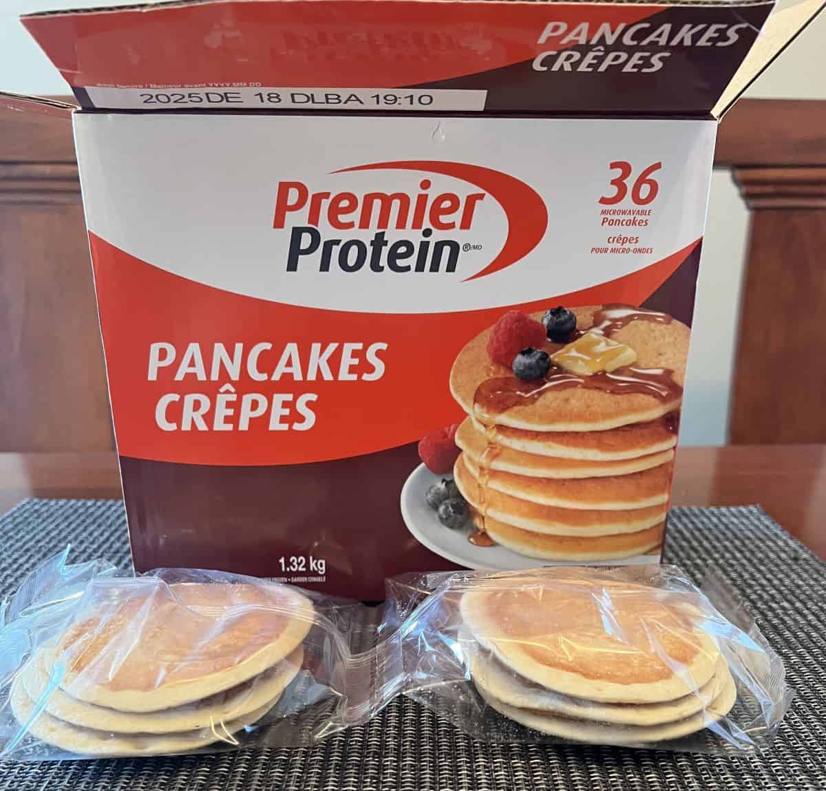 Image of the box of Premier Protein Pancakes open with two packs of three pancakes sitting in front of the box unopened.