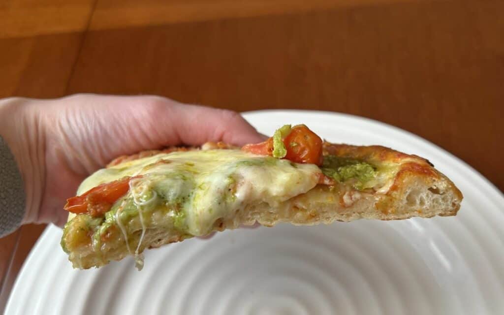 Closeup image of a hand holding one slice of Pinsa close to the camera so you can see how thick the slice it is. 