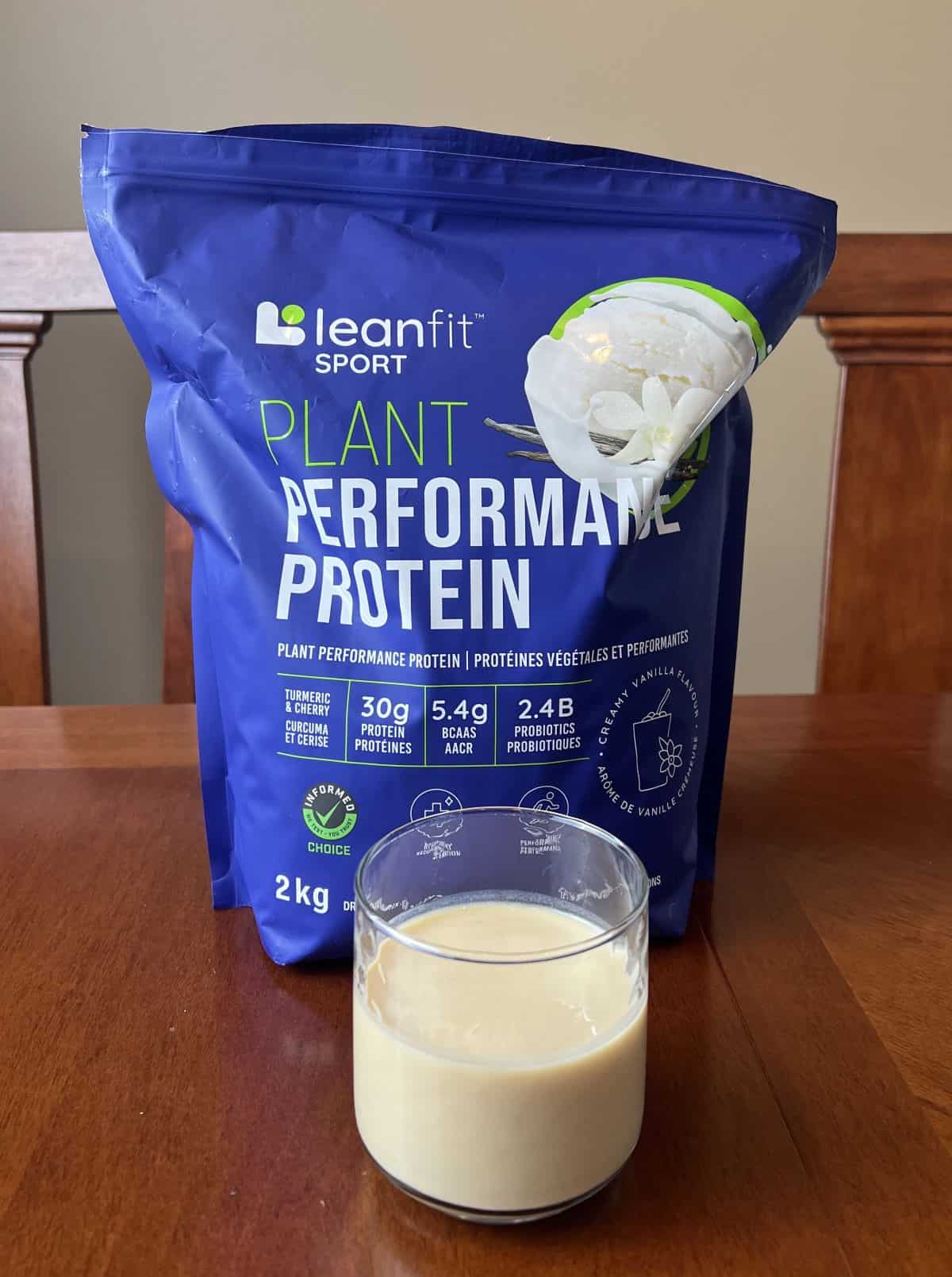 Image of the Costco Leanfit Sport Plant Performance Protein Review bag sitting on a table unopened with a glass with protein shake in it infront of the bag.