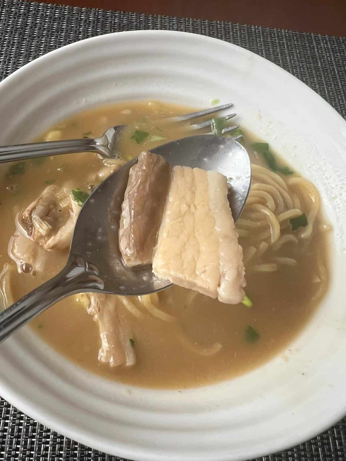 Image of a spoon hovering over a bowl of ramen, on the spoon is a piece of pork belly.