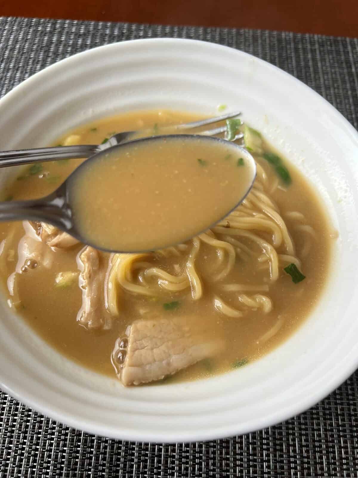 Closeup image of a spoon with ramen broth on it hovering over a bowl of ramen.