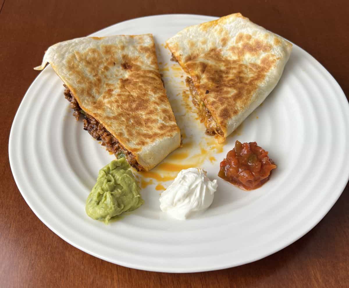 Image of two pieces of quesadilla served on a white plate with sour cream, salsa and guacamole on the plate.