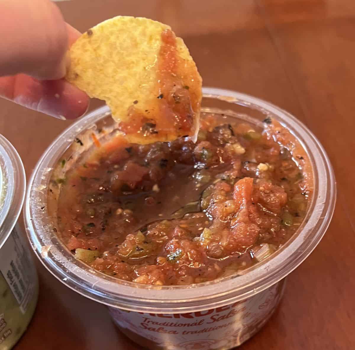 Image of a chip with salsa on it hovering over an open container of salsa.