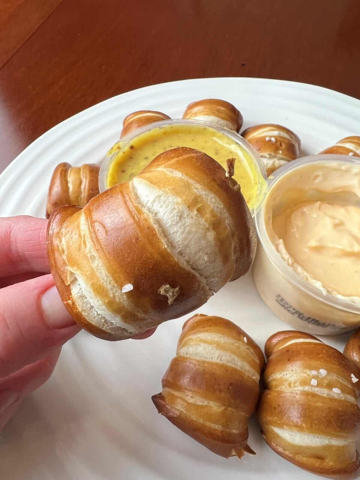 Image of a hand holding one pretzel bite close to the camera hovering over a honey mustard and cheese dip. 